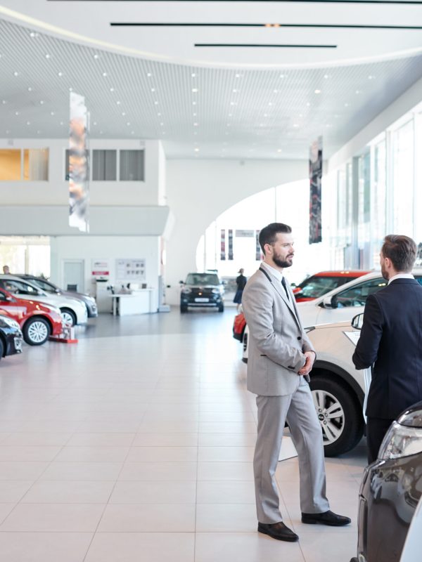 People in auto showroom Professional salesperson selling new cars in a modern auto salon. Two men standing and choosing a new car